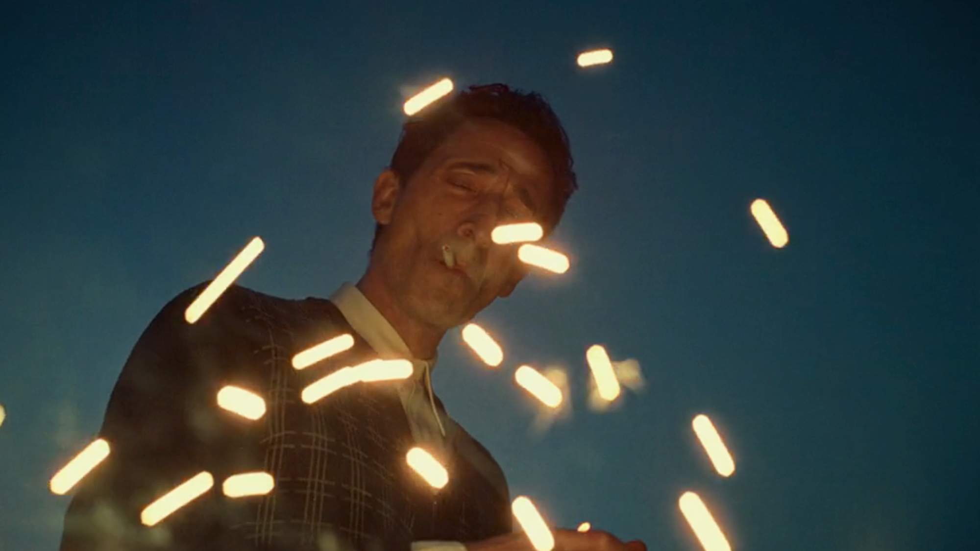 Adrien Brody smoking a cigarette with sparks flying in front of him in The Brutalist