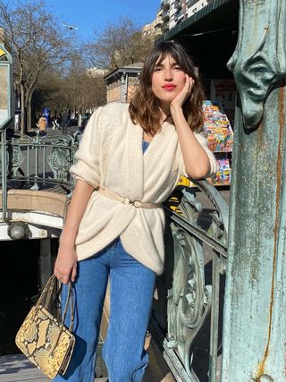 Jeanne Damas poses with a red lip, oversized white cardigan, and classic blue jeans