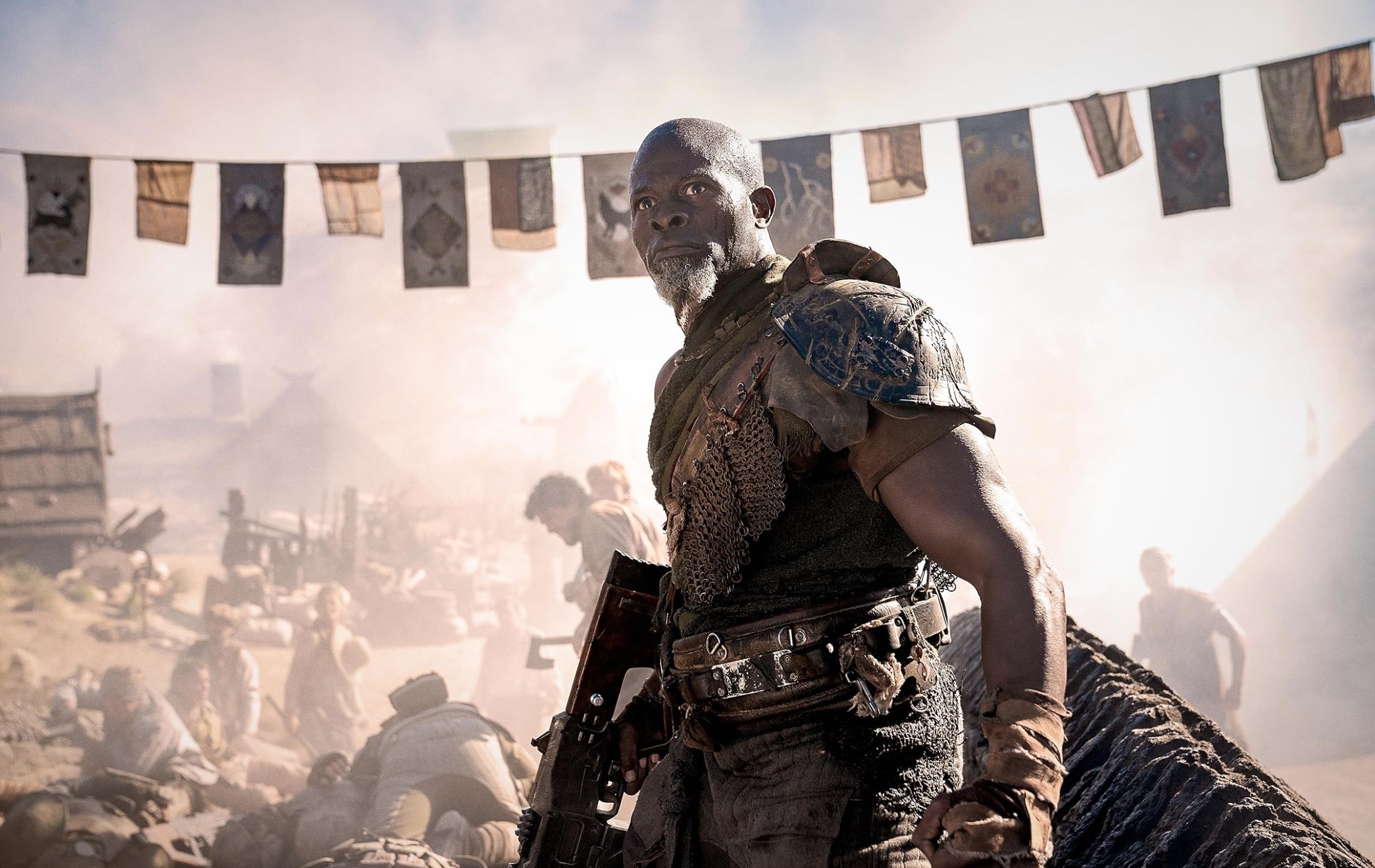 a man in gladiator-like armor stands ready to fight, with a chaotic scene in the background