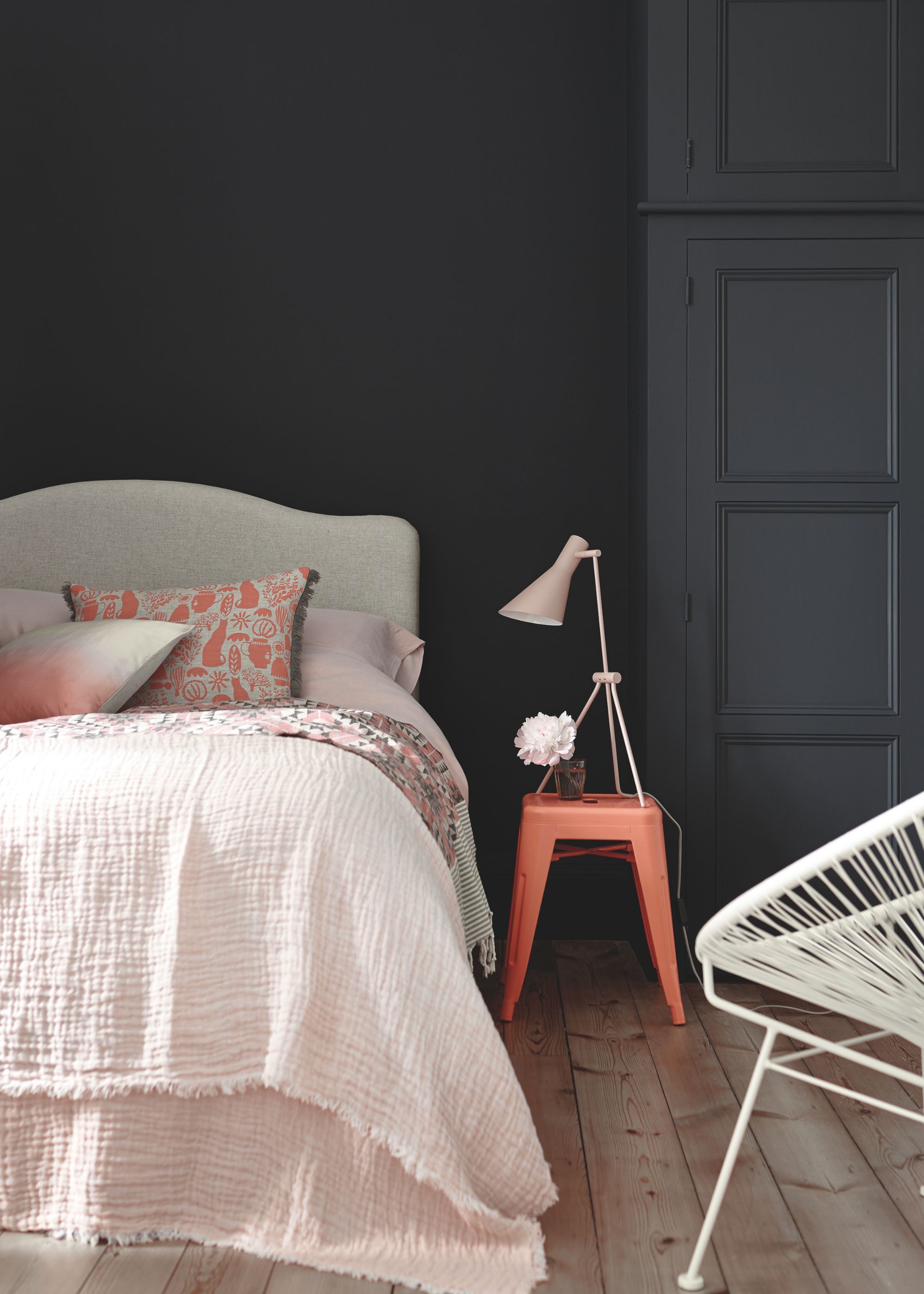 A bedroom with dark blue-black walls, a grey headboard with soft pink bedding, and a red nightstand with a white lamp.