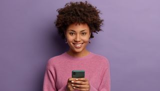 woman smiling holding phone wearing a pink sweater on a purple background