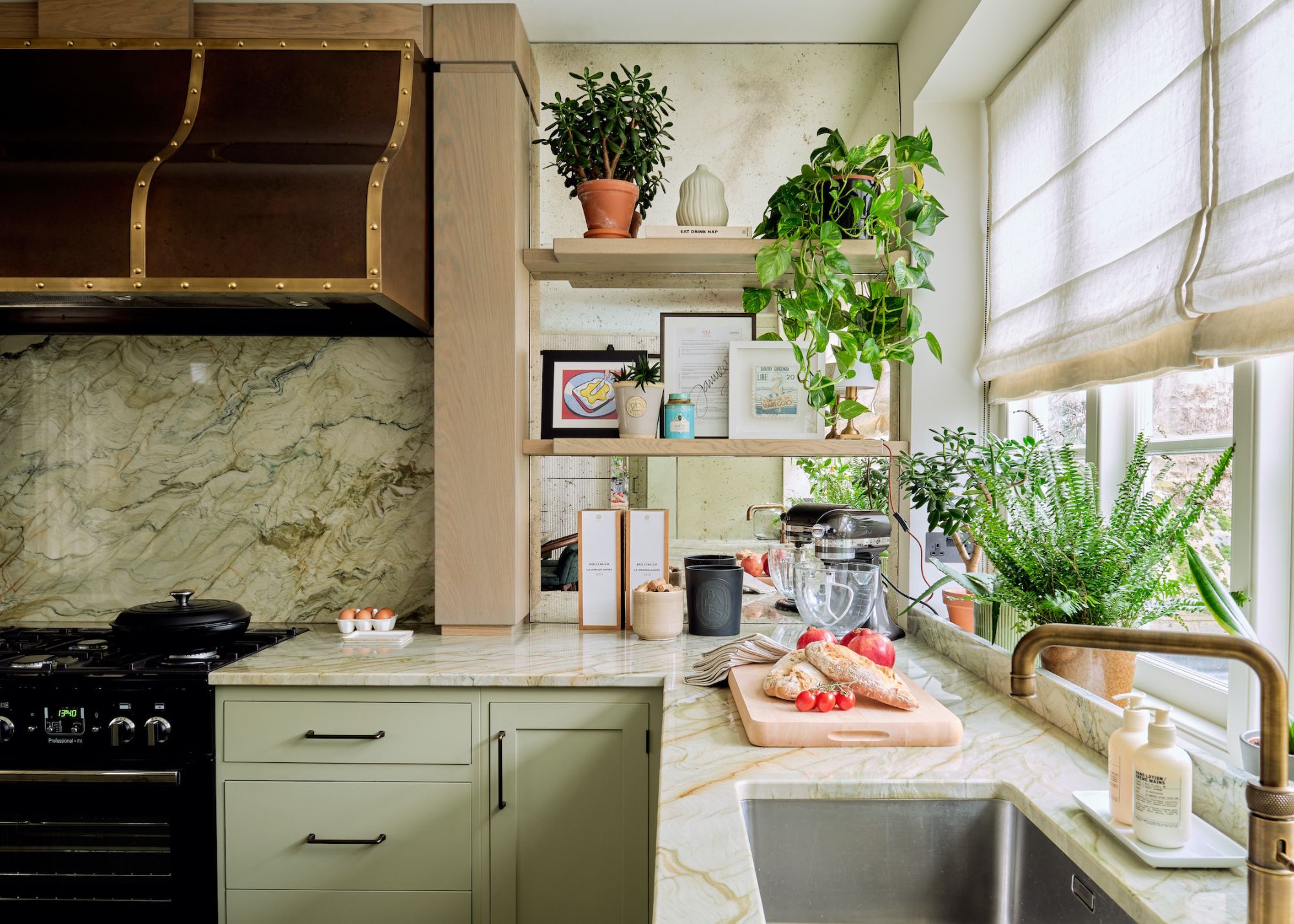 A kitchen with pale green cabinetry, a steel extractor hood, a black stove and oven, and a stone counter with a mirrored shelving nook featuring potted plants and herbs alongside print and photo frames and cooking accessories by a stand mixer and a chopping board with bread and tomatoes by a sink with a tray of Le Labo soaps