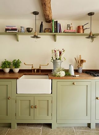 A sage green country kitchen in a Cotswold stone cottage with Belfast sink, open shelving, shaker kitchen cabinets with brass knobs and brass tap