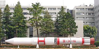 A white and red rocket at the bottom of the screen. A building in the background is mostly covered by trees.