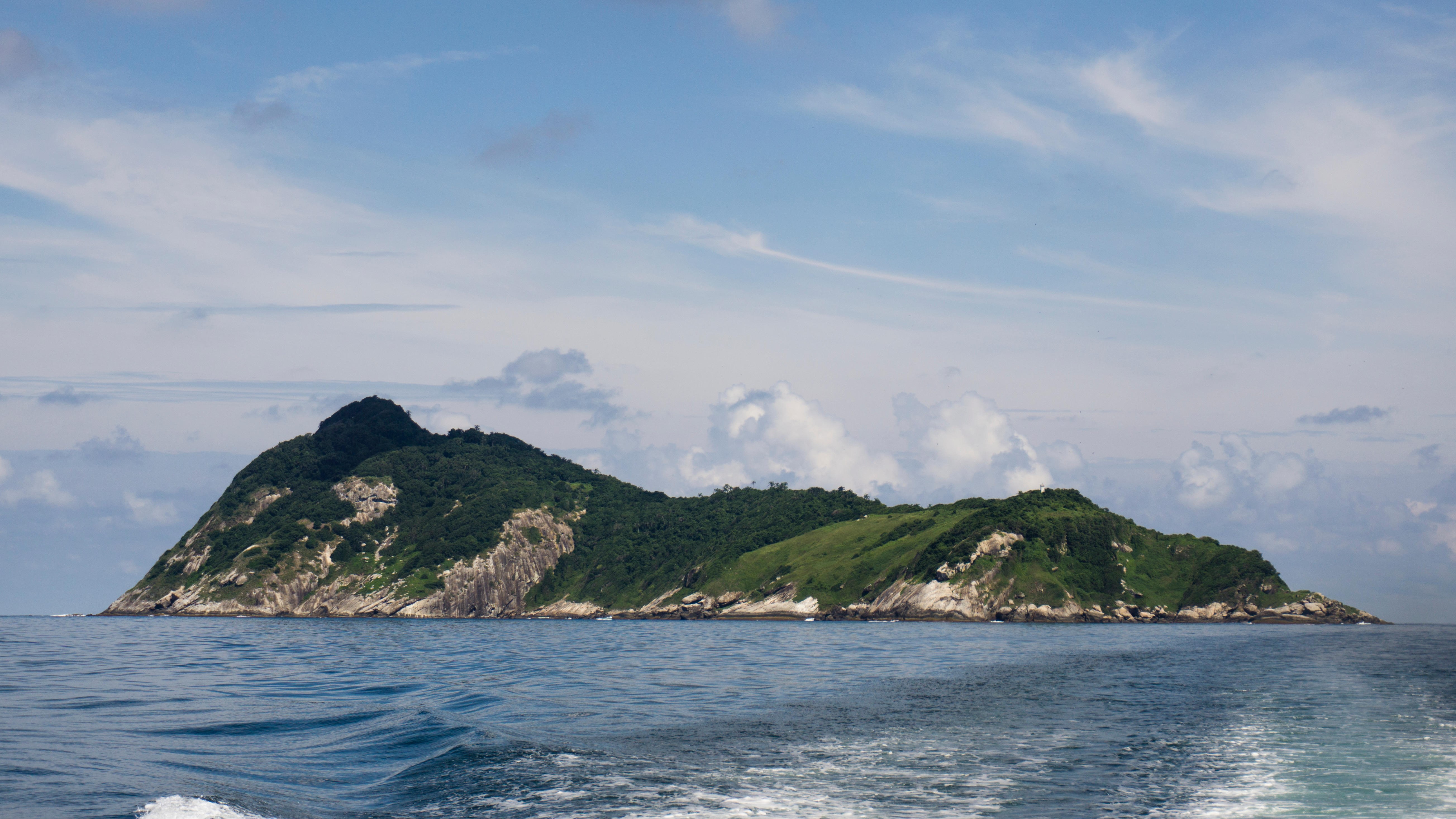 Uma foto da Ilha das Cobras tirada de um barco no Oceano Atlântico. A ilha tem um terreno variável de florestas, pastagens e rochas áridas.