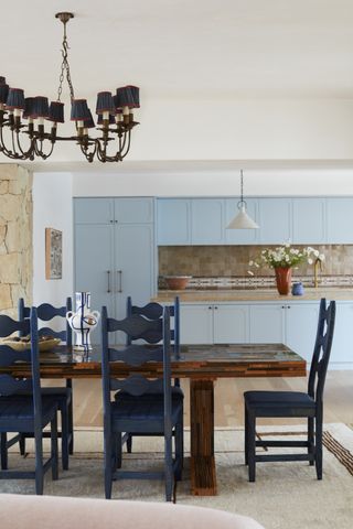 light blue kitchen with tiled backsplash, with a timber dining table in foreground with stained navy blue chairs, a chandelier, and a beige rug underneath