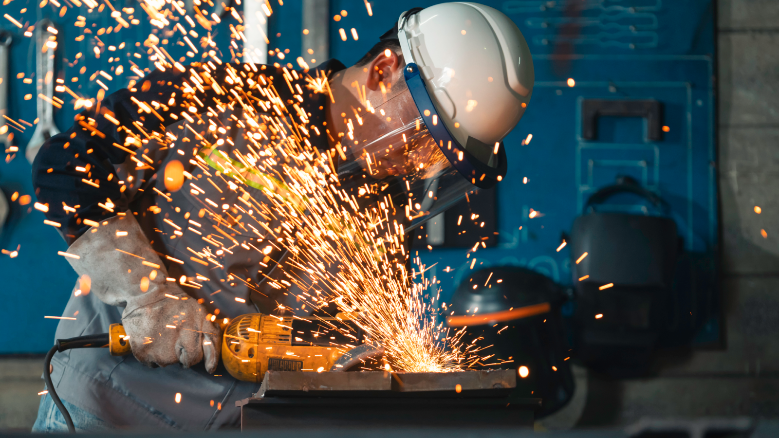 A construction worker in safety helmet and gloves uses an angle grinder on a steel plate, sending a cascade of bright orange sparks across a dim industrial workshop.
