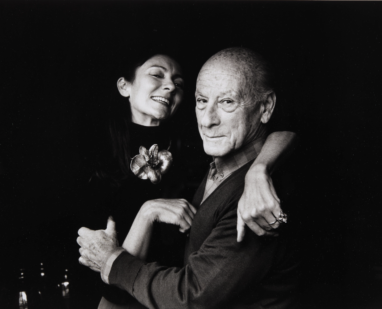 Pauline Karpidas and her husband Constantinos in a black and white photo. Pauline wears a large flower and a highneck top, they are embracing
