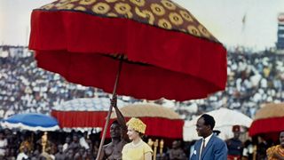 Queen Elizabeth II makes her way underneath a large, gaily colored umbrella, to a dais to watch the Durbar of the Ashanti Chiefs, at Kumasi Sports Stadium.