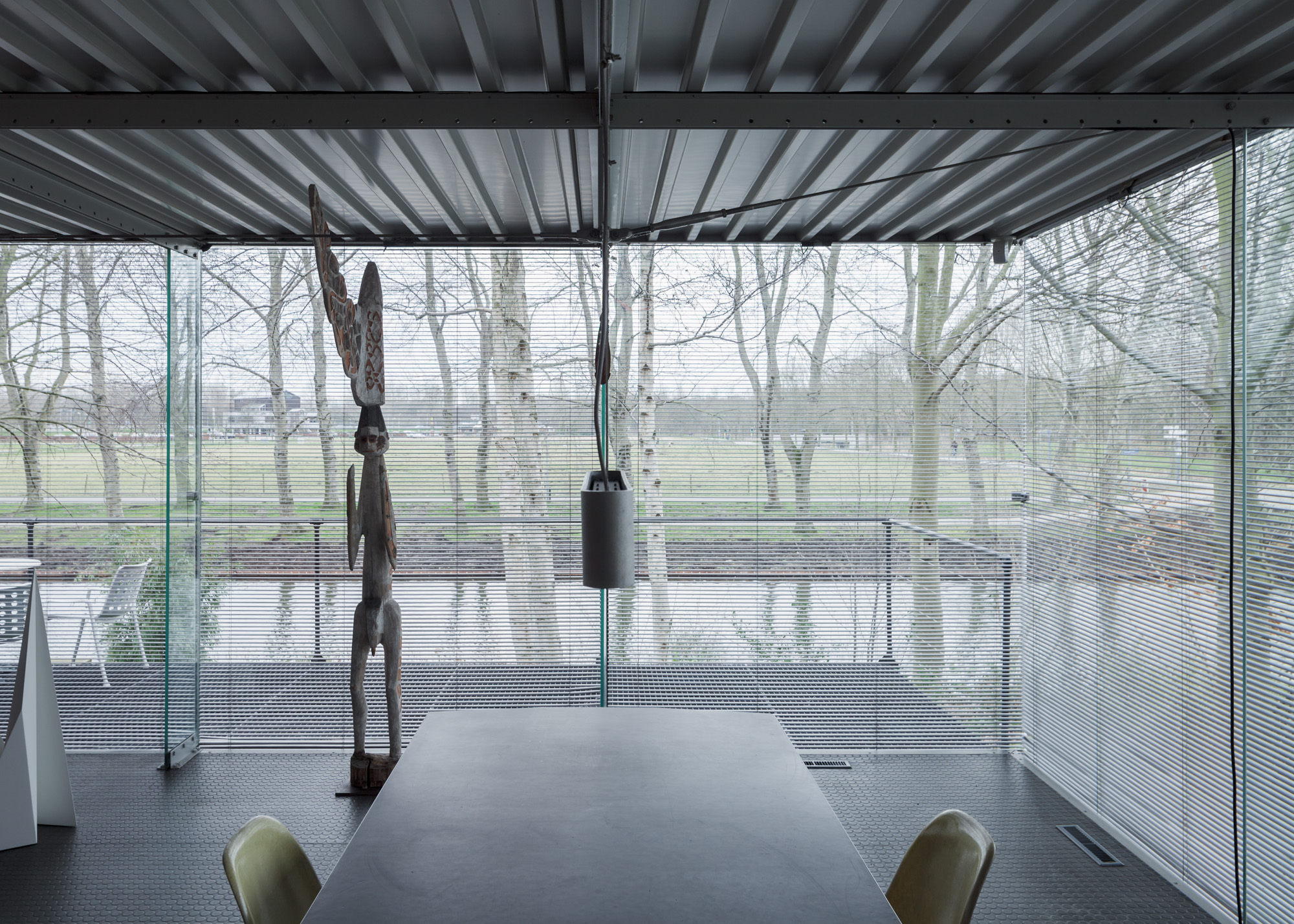 view out of the house onto a landscape with bare trees through floor-to-ceiling glass windows. a wooden sculpture on the left