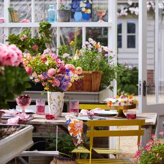 greenhouse with table and chairs set up for entertaining, plants, flowers, cake, pink tumblers, fruit, artwork