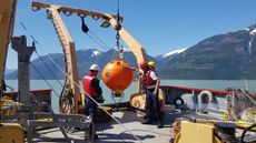 Three men stand on the deck of a ship working with a large ball-shaped instrument suspended in the air