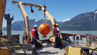 Three men stand on the deck of a ship working with a large ball-shaped instrument suspended in the air