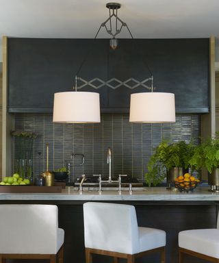 A kitchen with a steel black pendant light with two lampshades, a gray kitchen island with a silver faucet and gray bar stool chairs in front of it, and a dark gray splashback and oven behind it