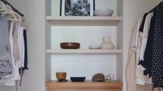 A walk-in wardrobe with an open rail system and an in-built storage nook in the middle with ceramics, a cap, and a leaning frame