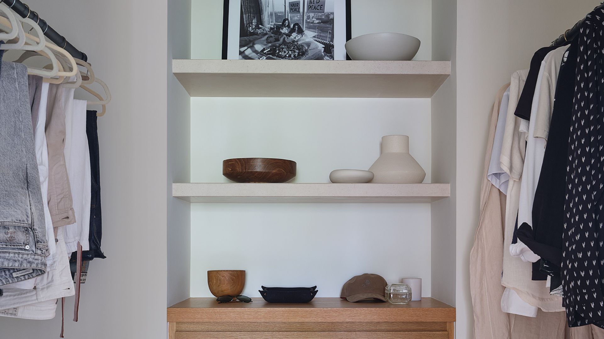 A walk-in wardrobe with an open rail system and an in-built storage nook in the middle with ceramics, a cap, and a leaning frame