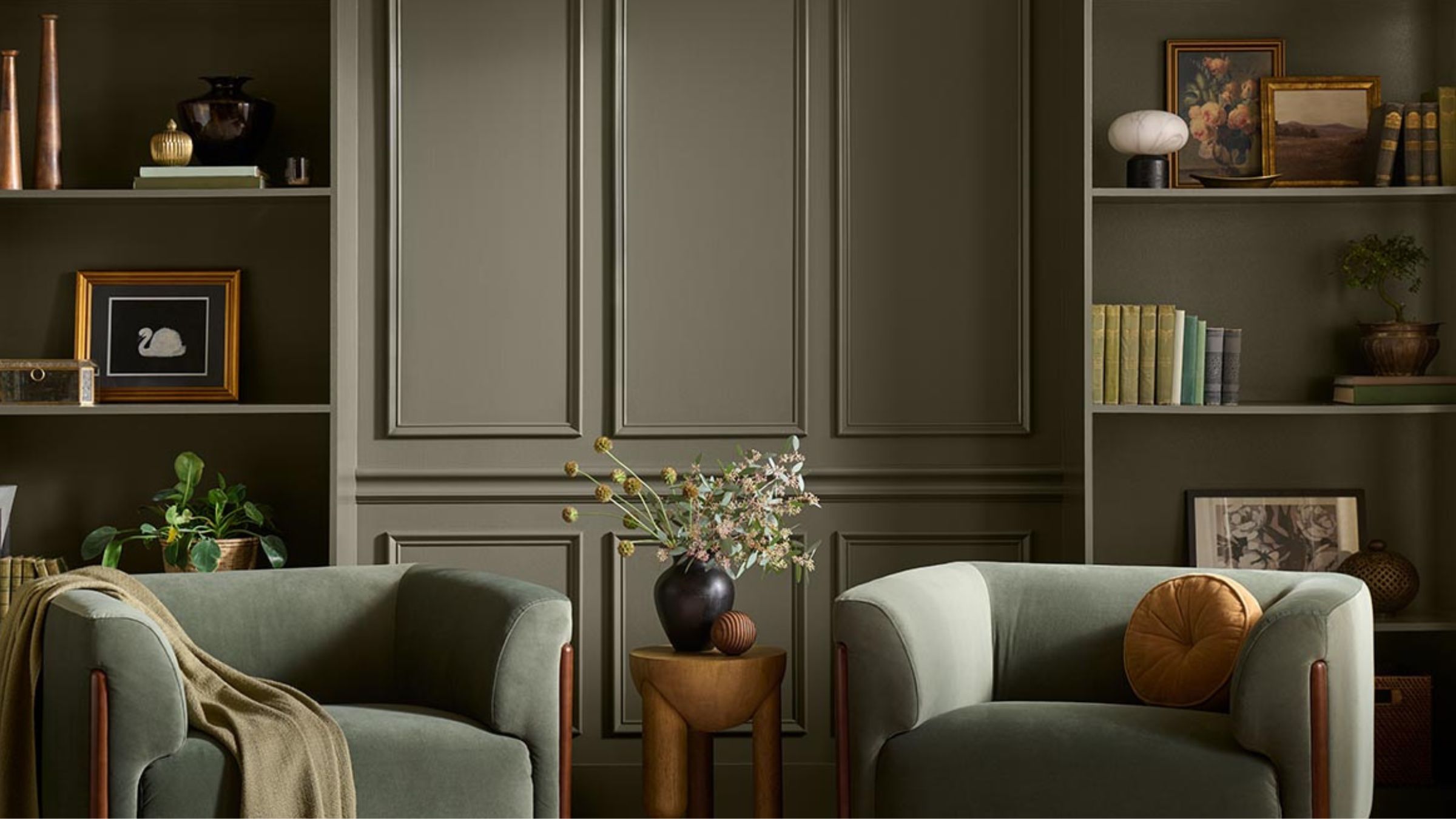 Living room bookcases and wall painted in Behr's Grounded, with two velvet green armchairs, a side table with a vase of flowers, and bookshelves covered in decor.