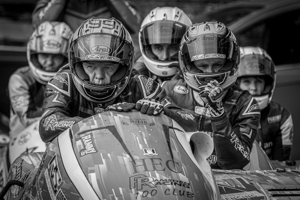 A group of motorcyclists in racing gear pose together in a monochrome image, showcasing their helmets and a colorful racing machine