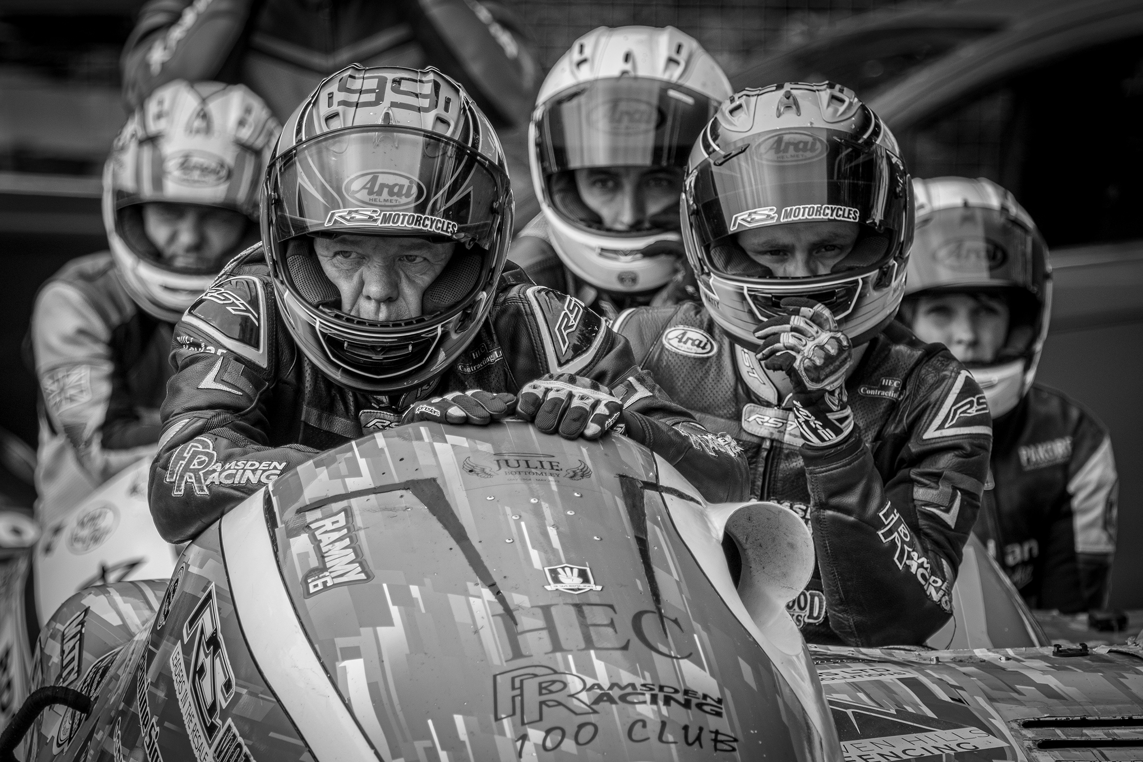 A group of motorcyclists in racing gear pose together in a monochrome image, showcasing their helmets and a colorful racing machine