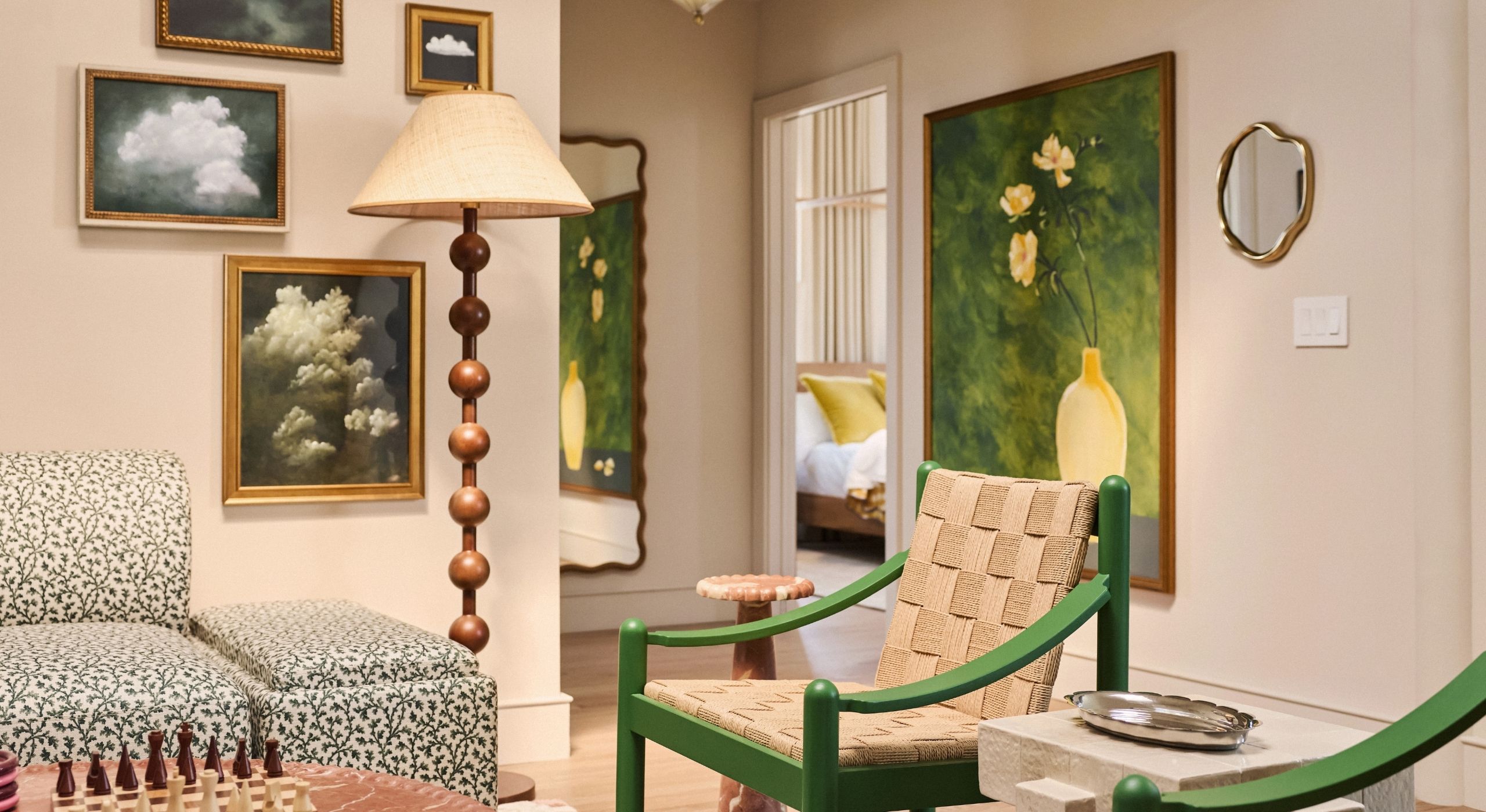 A creatively decorated suite with framed artworks, an eye-catching upholstered couch in black and white, floral patterns, rattan and wood beige and green chairs, and a tall, standing bobbin lamp.