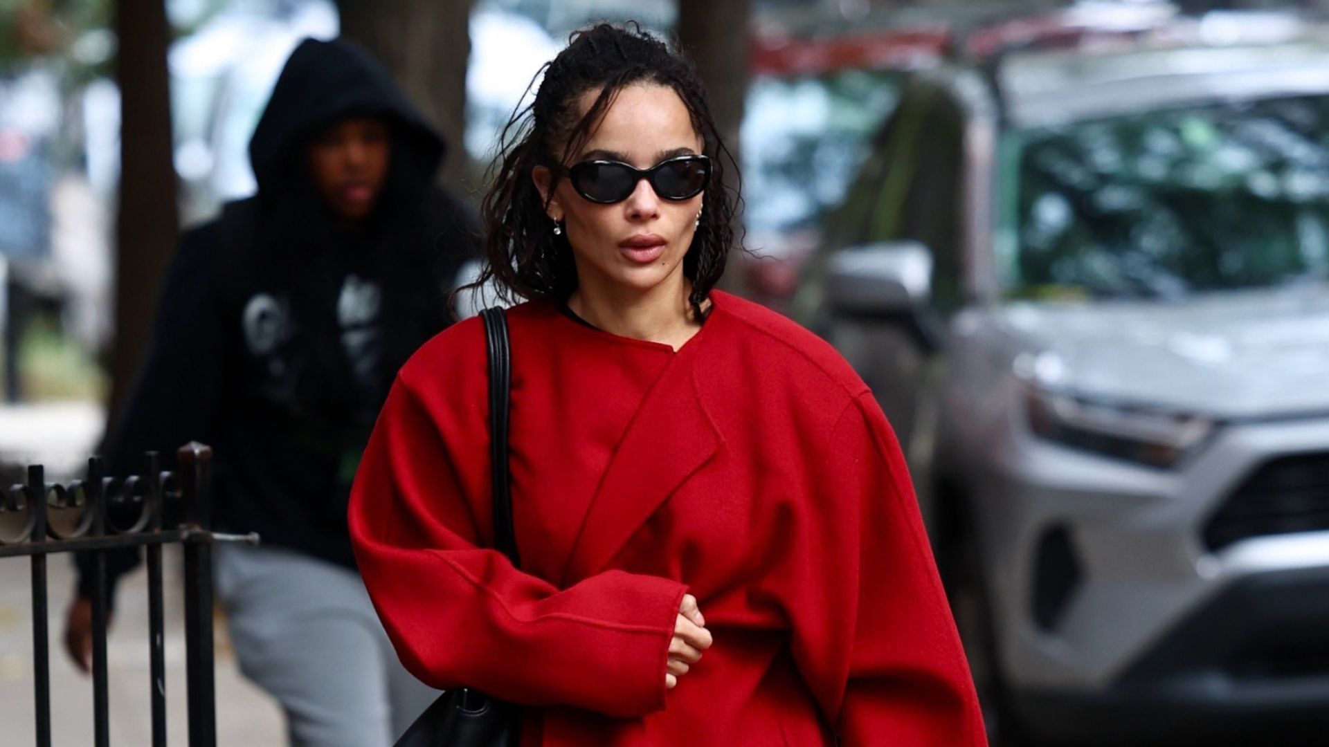 Zoe Kravitz walks down the street in New York wearing a red collarless coat with black trainers, a black leather bag and black sunglasses. She holds a transparent shopping bag and a brown paper bag.