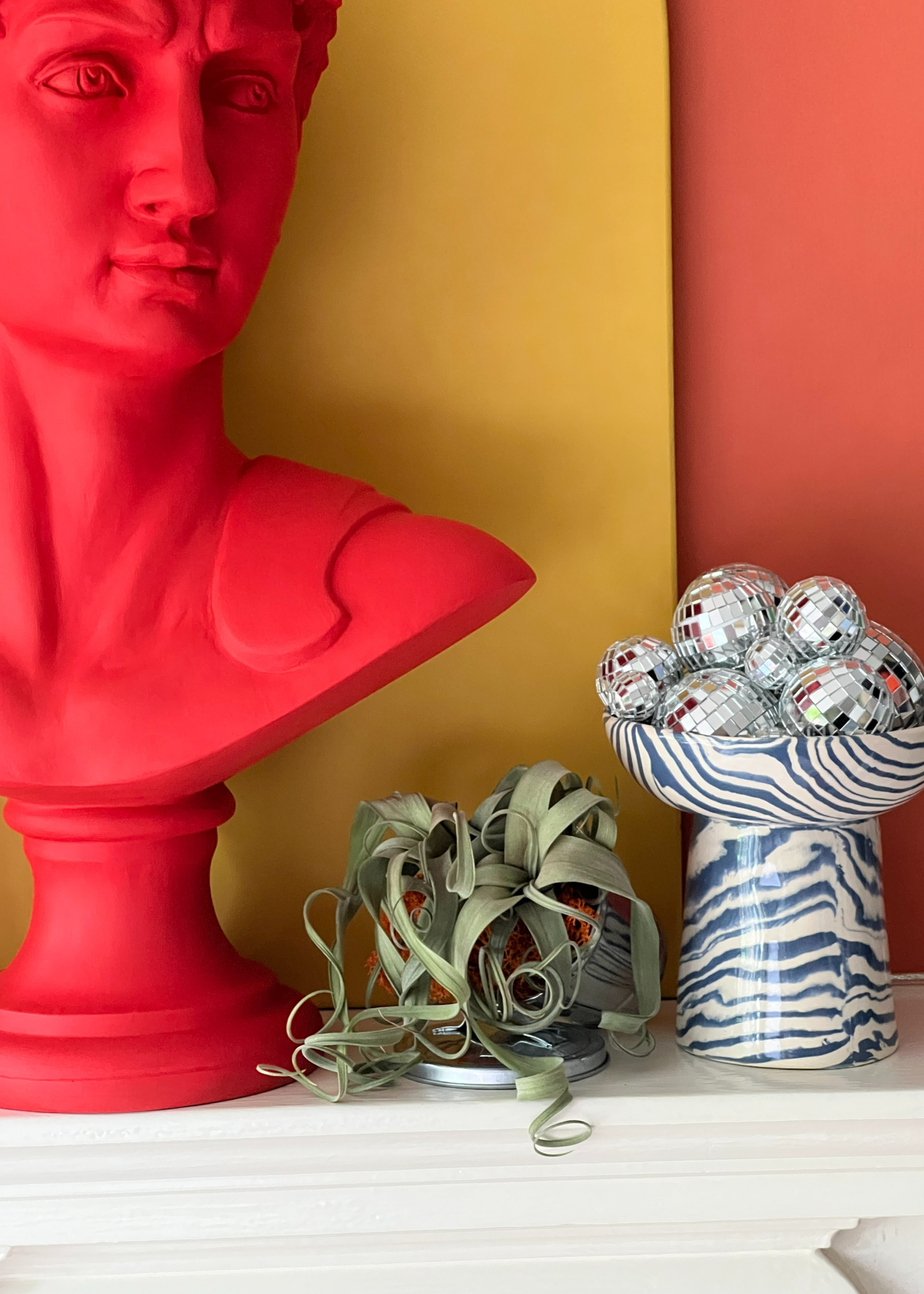 A mantel with a bold red bust, a potted plant and a pedestal bowl with mini disco balls