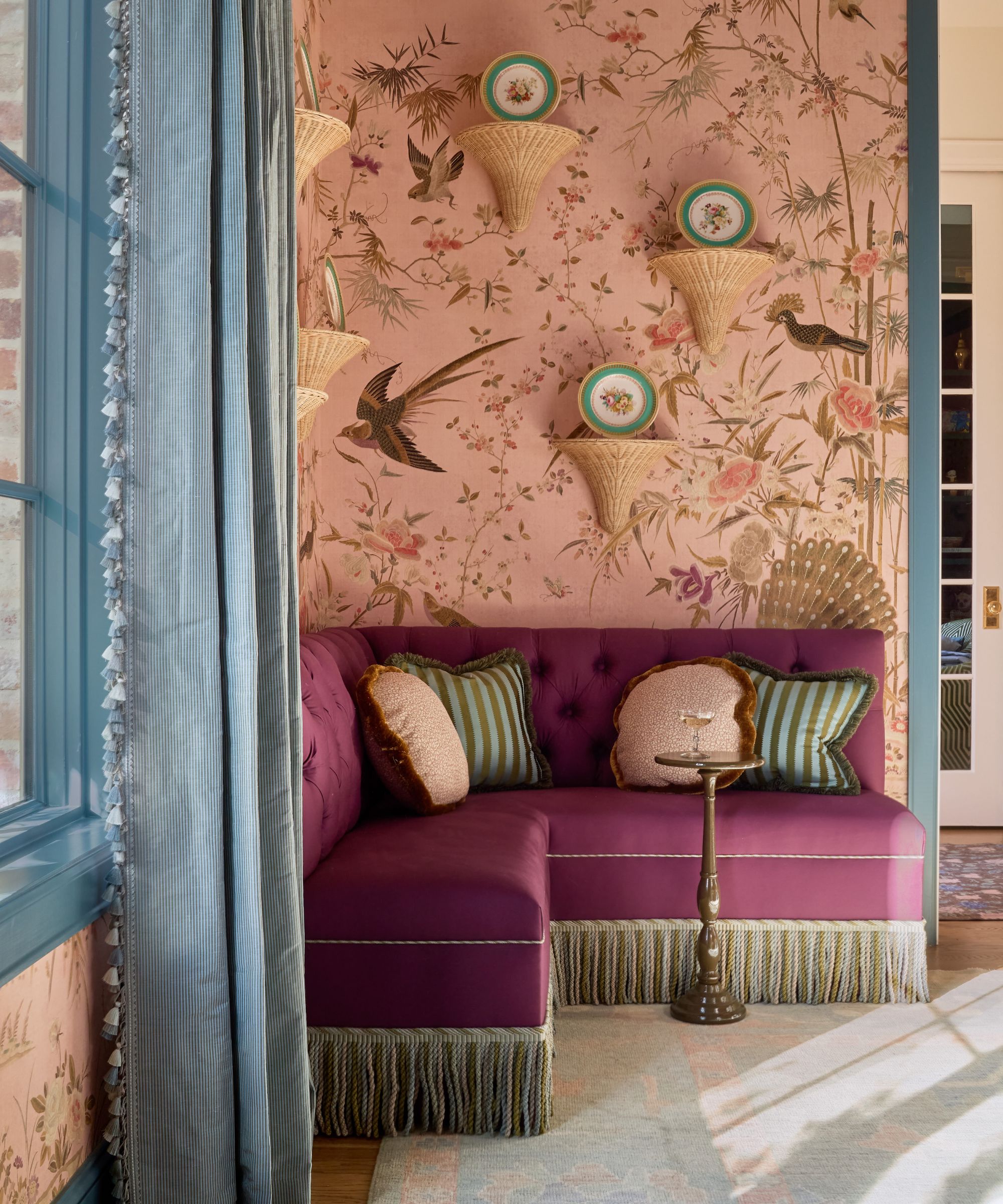 Cozy corner featuring a plush purple velvet fringed banquette, striped cushions, pink chinoiserie wallpaper, and blue tassel-trimmed drapes.