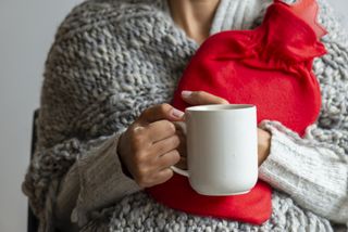 A woman clutching a red, hot water bottle close to her chest, with a white mug in her other hand.