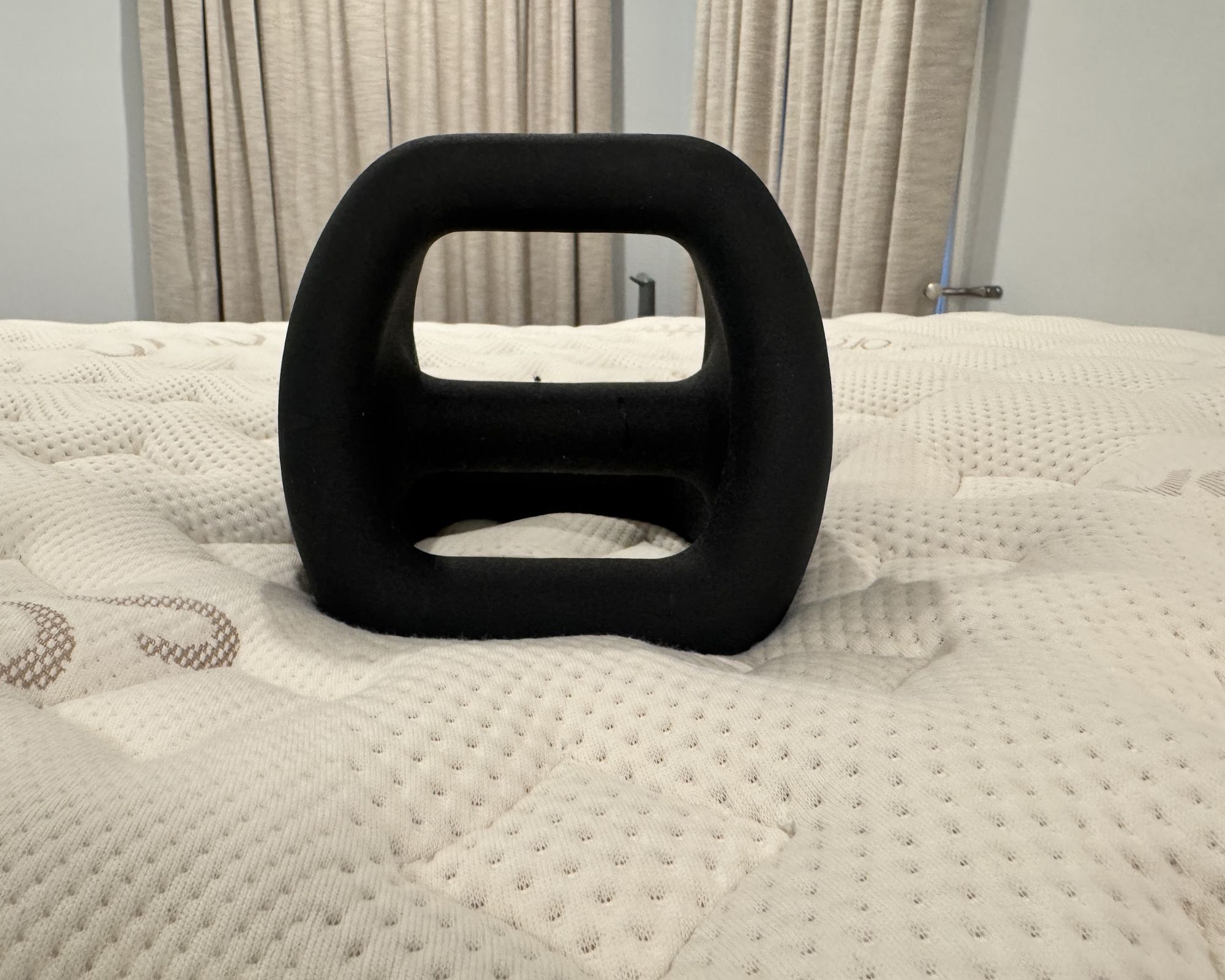 A black weight pictured on top of the Saatva RX mattress, with tan curtains and pale blue walls behind.