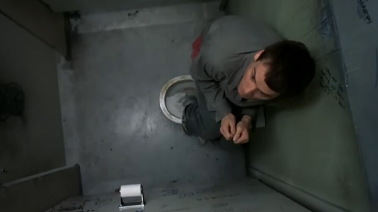 Lloyd Christmas (Jim Carrey) hides in a bathroom stall, standing on a toilet.