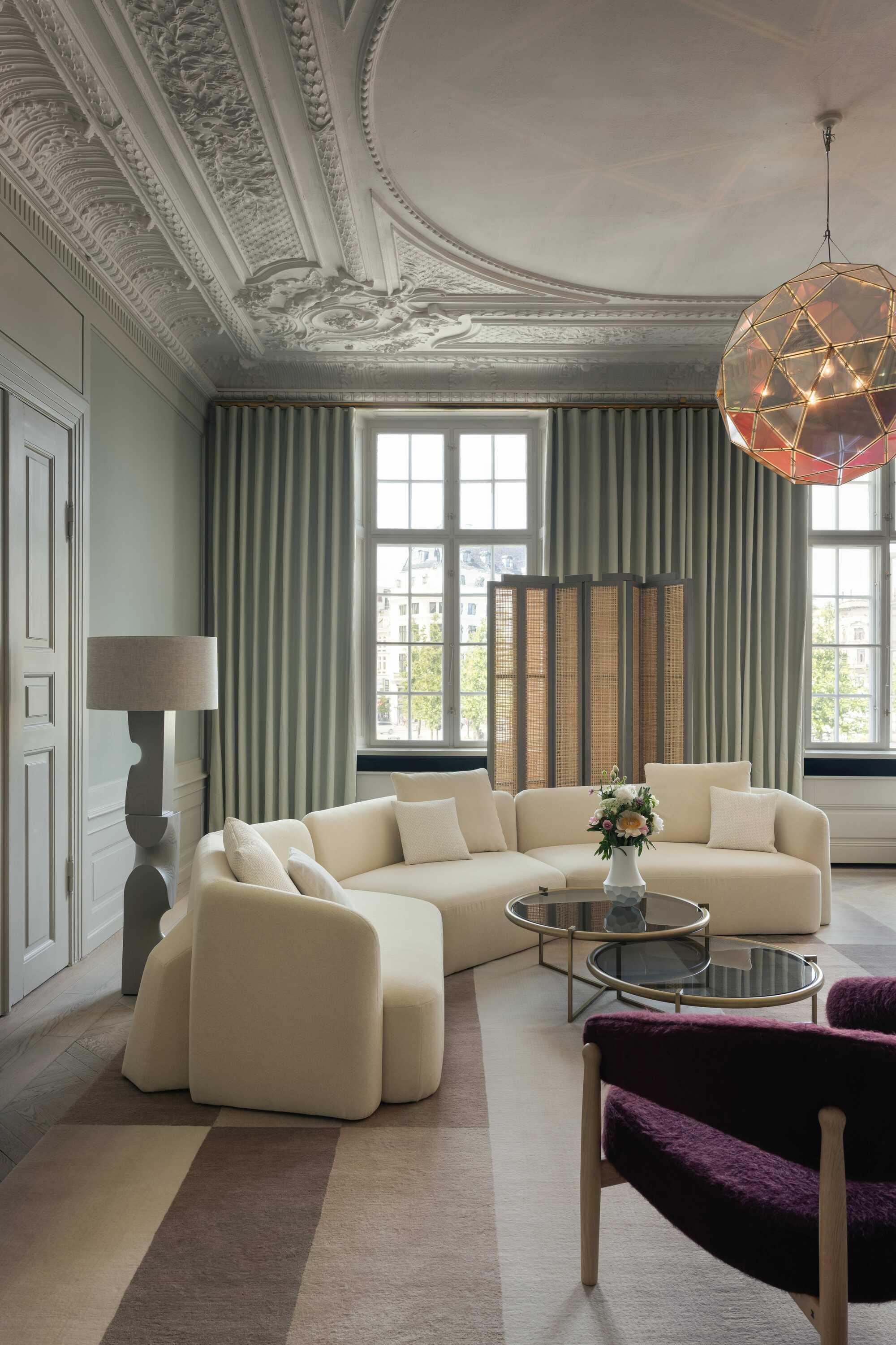 white boxy curved sofa in a classical living room with ornate cornicing, curtains, striped rugs, purple armchair, floor lamp and room divider with rattan