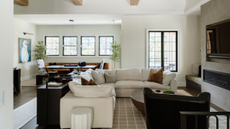 an inviting living room with a large cream sectional, a leather accent chair, a patterned rug, pillows, a dark console table behind the couch holding decor, and a TV built into the wall above a fireplace 