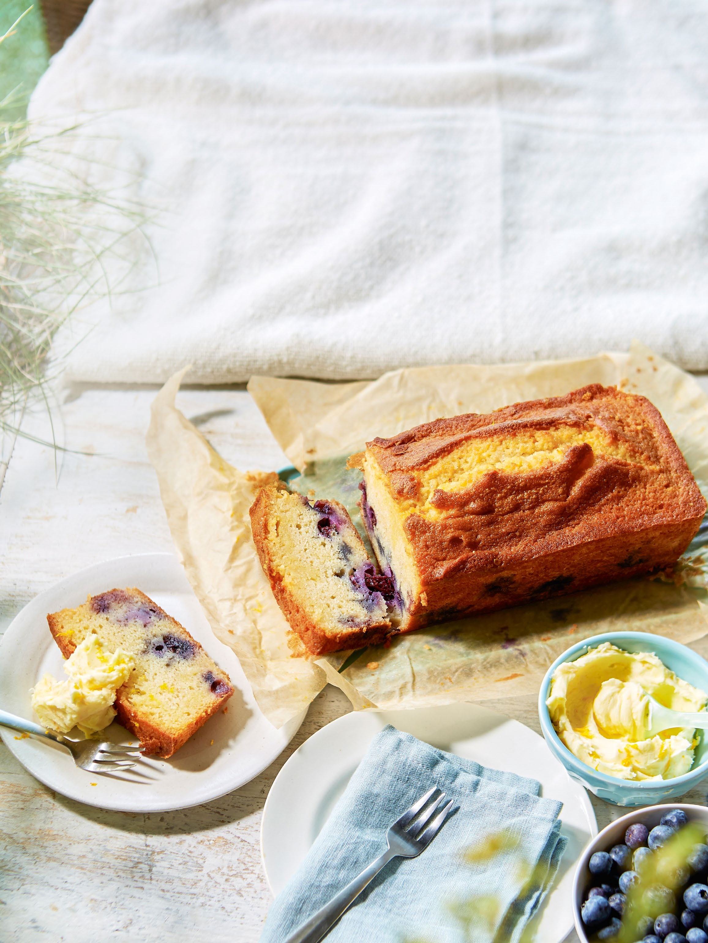 A freshly baked blueberry loaf cake on parchment paper, with a slice served on a plate alongside butter, set against a light background, perfect for a picnic.