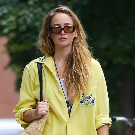 Jennifer Lawrence wearing a yellow Bode button-down shirt with sunglasses, a white tank top, white trousers, a Loewe raffia tote, and Melissa jelly shoes.