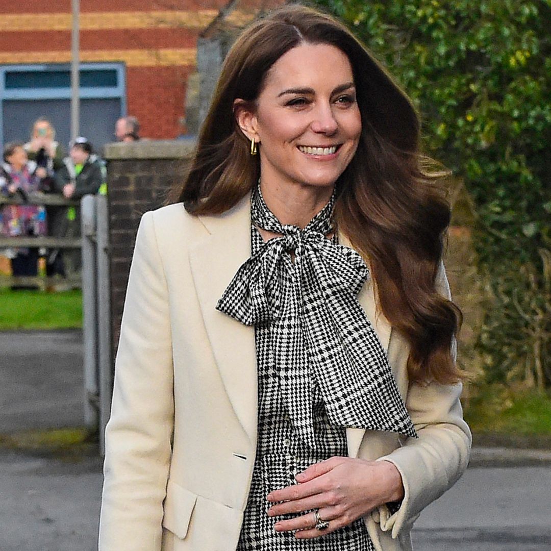 Kate Middleton wearing a black checkered dress and cream coat smiling walking in front of a wall with green leaves