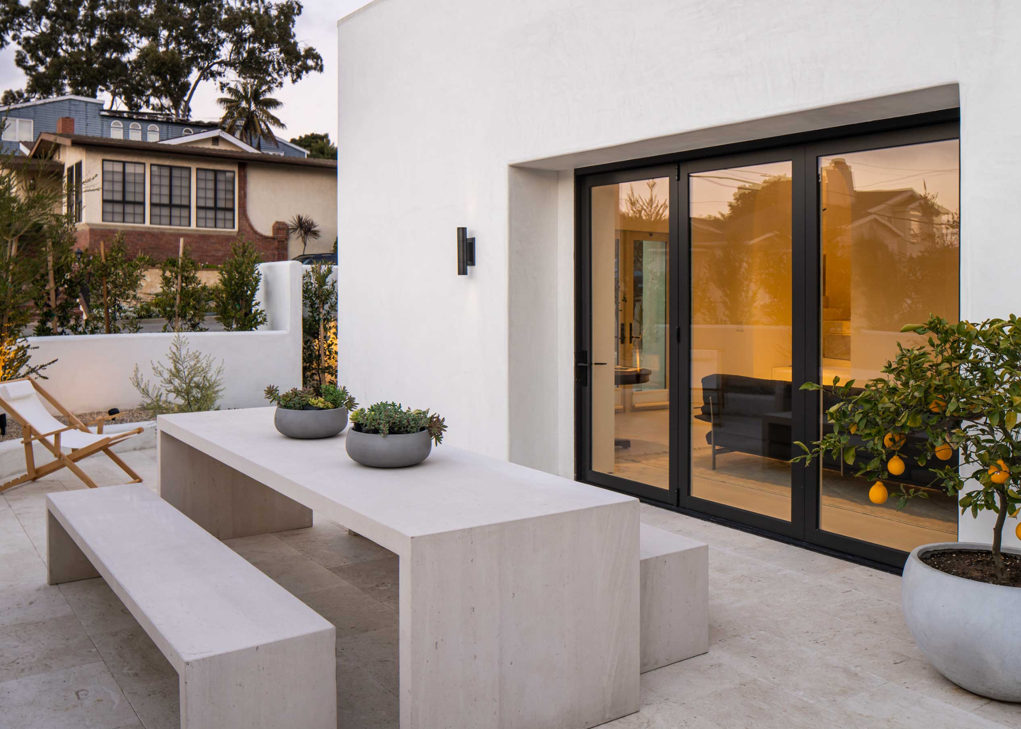 a minimalist patio in a backyard with a table in the middle as well as benches on each side. There is also a plant with oranges hanging off of it