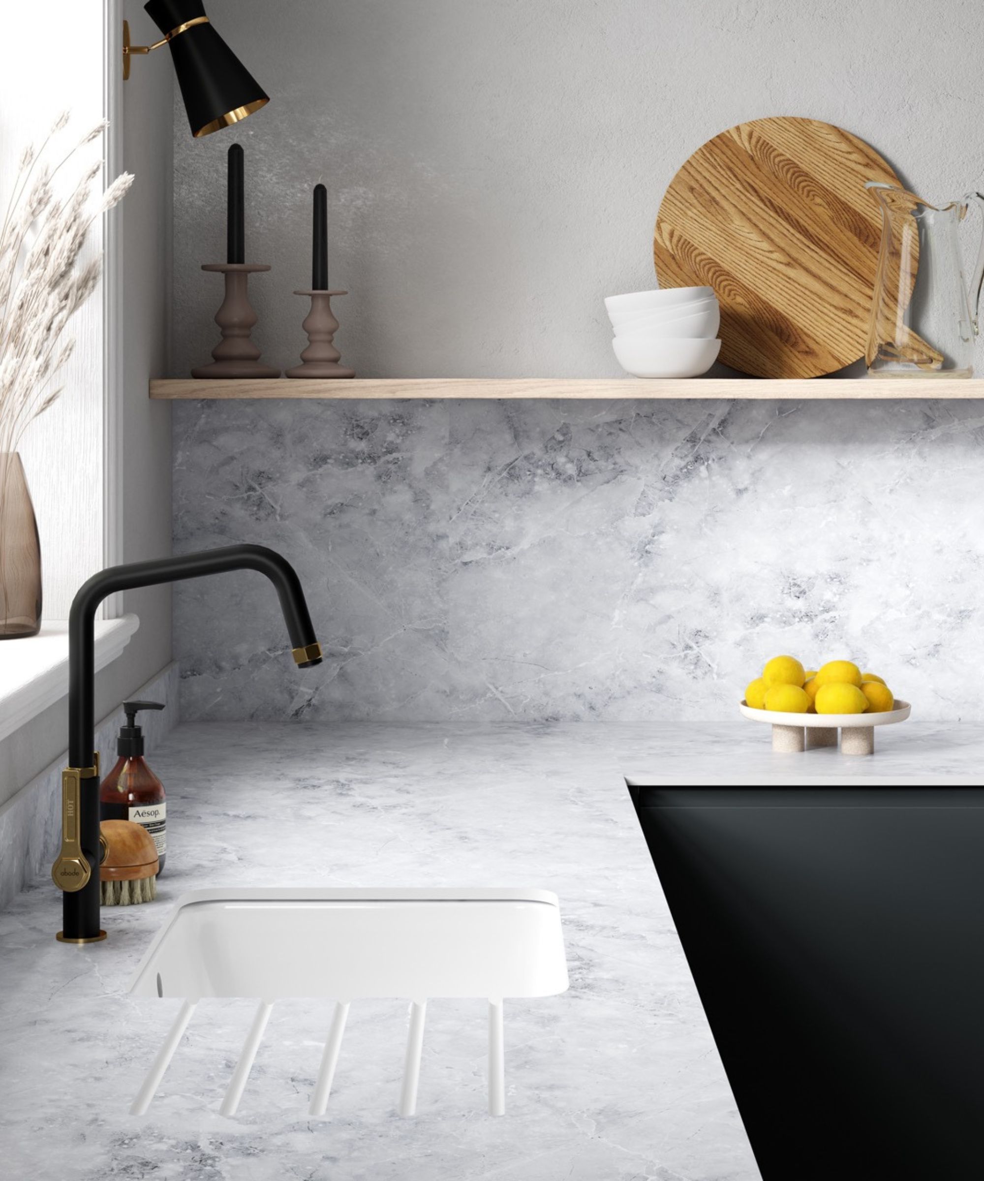 Contemporary kitchen sink area with pale marble-effect worktops, a white inset sink and matte black tap with brass detailing. Open wooden shelving above holds ceramics and chopping boards, styled with neutral accessories and lemons in a bowl.