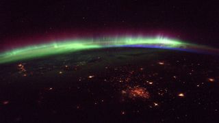A photo of a green, blue and purple aurora over the horizon of the earth, with reddish city lights in the foreground