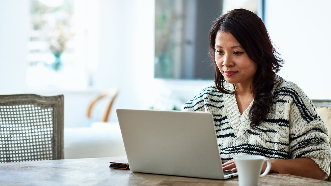 A woman sits at her dining room table and looks at her laptop.