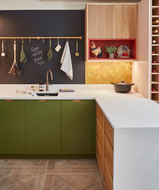 A modern kitchen with deep green lower cabinets, white countertops, and a black chalkboard wall with a brass rail holding kitchen essentials. A rooster figurine sits in a red shelf nook, and a wine rack is built into the cabinetry.