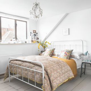 A bedroom with a double bed and paisley patterned quilt, venetian blinds and chandelier, dormer window and wood floor