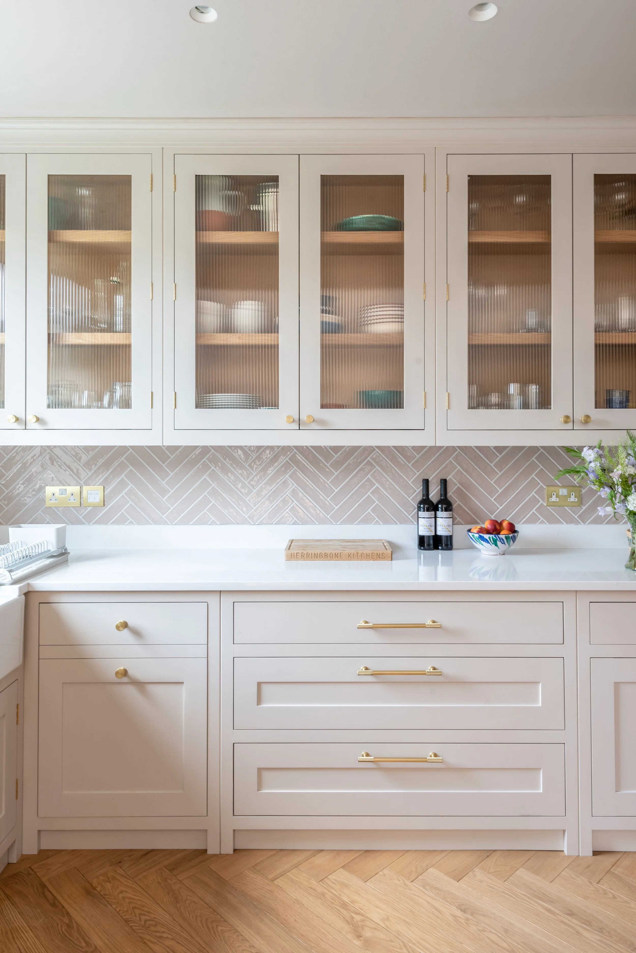 A blush pink kitchen with a row of reeded glass wall cabinets and white worktops. shown with herringbone tiled splashback and herringbone timber floors
