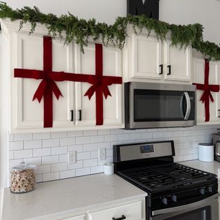 A kitchen with white cupboards wrapped in red velvet ribbon with bows on top and finished with a Christmas garland on top of the cabinets