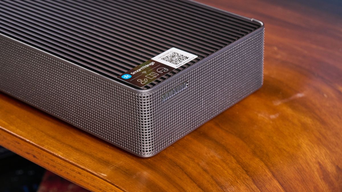 the samsung qs750f soundbar with two rear surround speakers and subwoofer photographed against a blue background and a TV