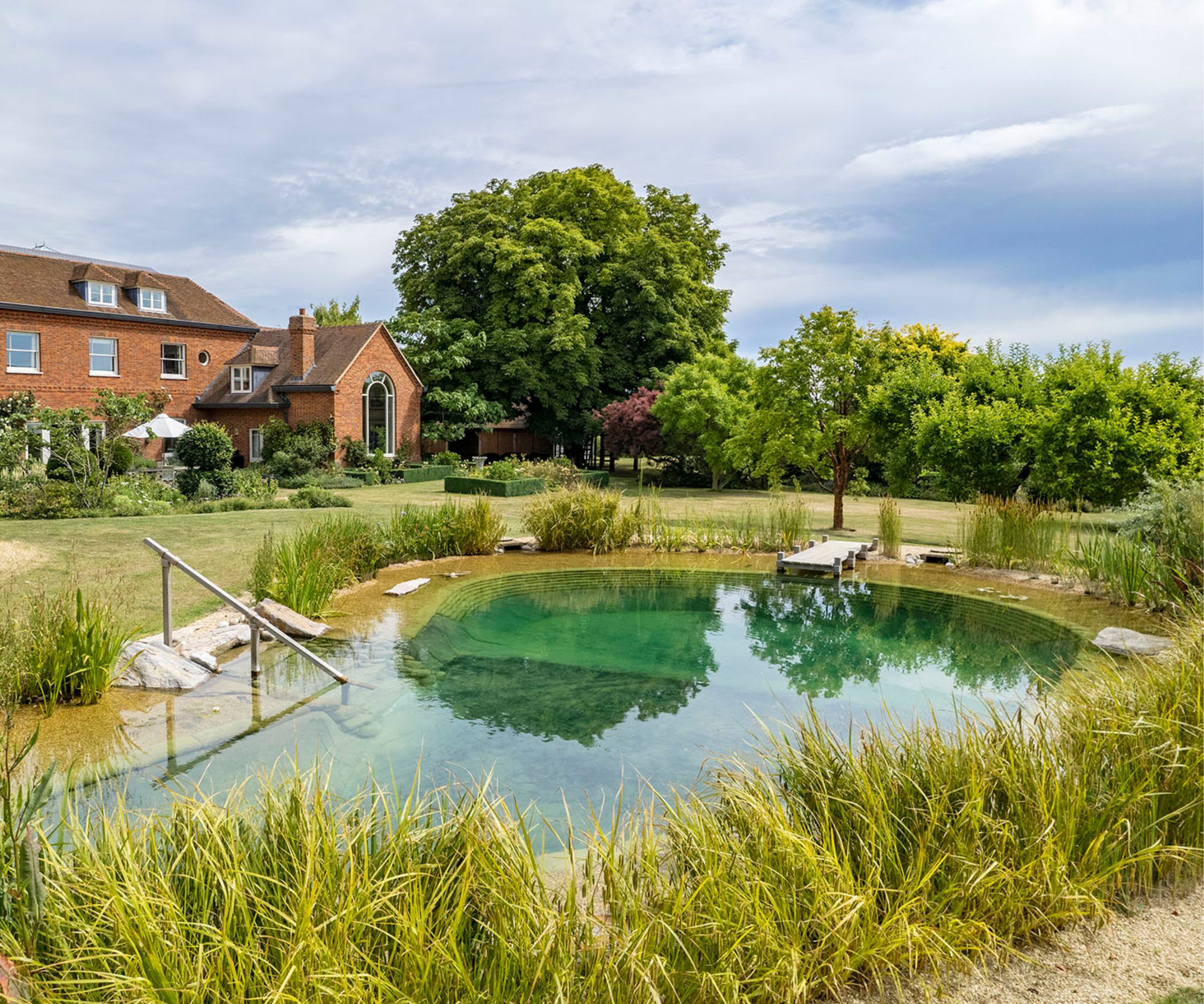 Beautiful natural swimming pool in a landscaped garden beside a large red-brick home, set against a backdrop of trees and open sky.