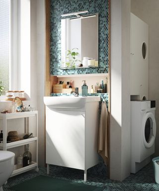 Small bathroom with white vanity, washer and dryer behind in alcove above each other, storage units, green blue mosaic tiles
