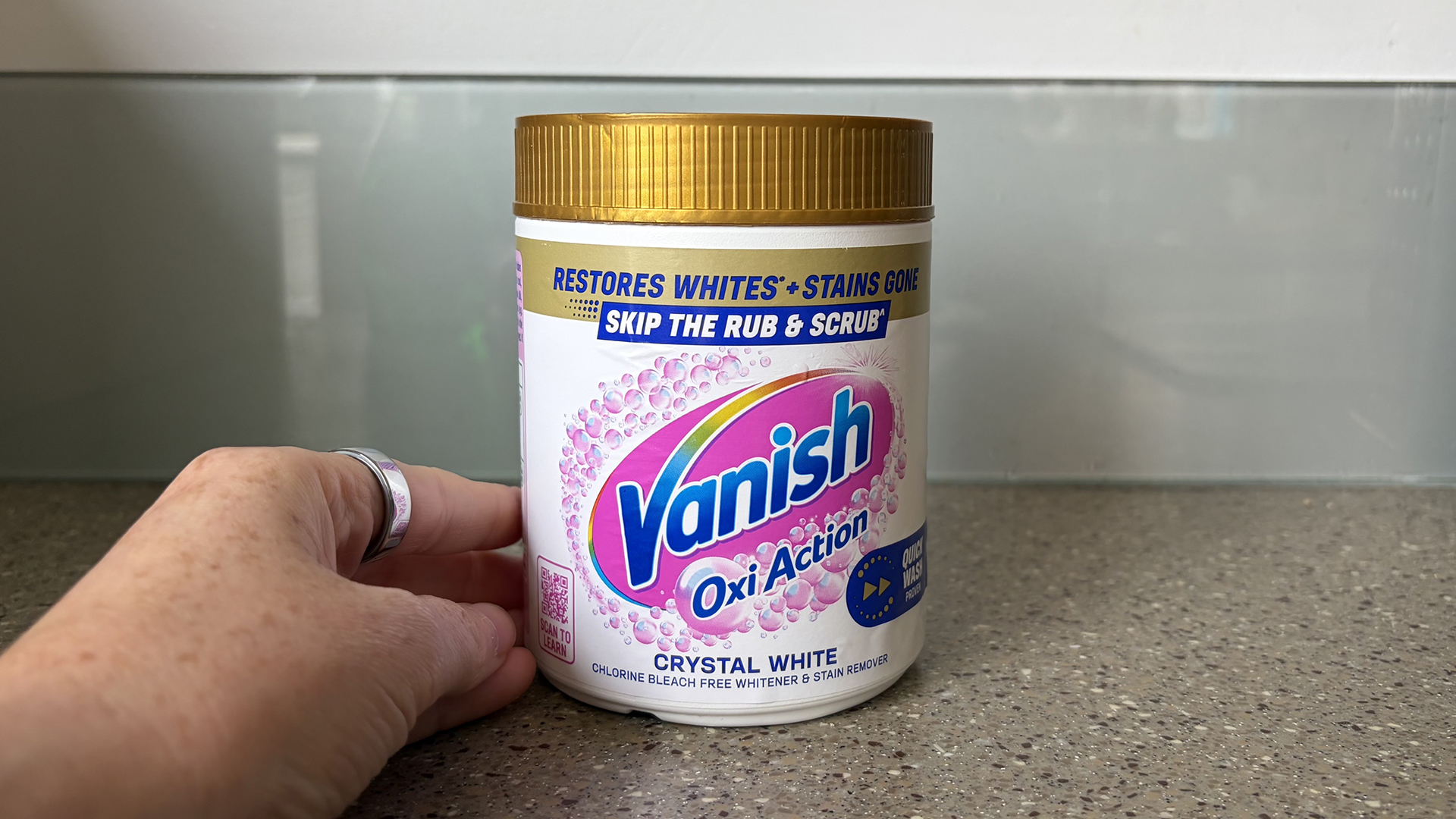 A close up of a hand next to a tub of Vanish OxiAction Oxygen Bleach on a kitchen counter