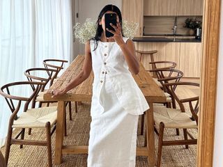 British fashion influencer Monikh Dale poses for a mirror selfie in front of her chic rustic wooden dining table and wishbone chairs wearing a white linen set with a long vest and maxi skirt