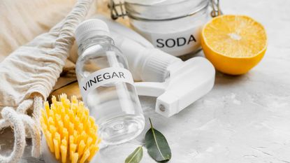 Baking soda and vinegar in glass jars beside a scrubbing brush and a halved lemon