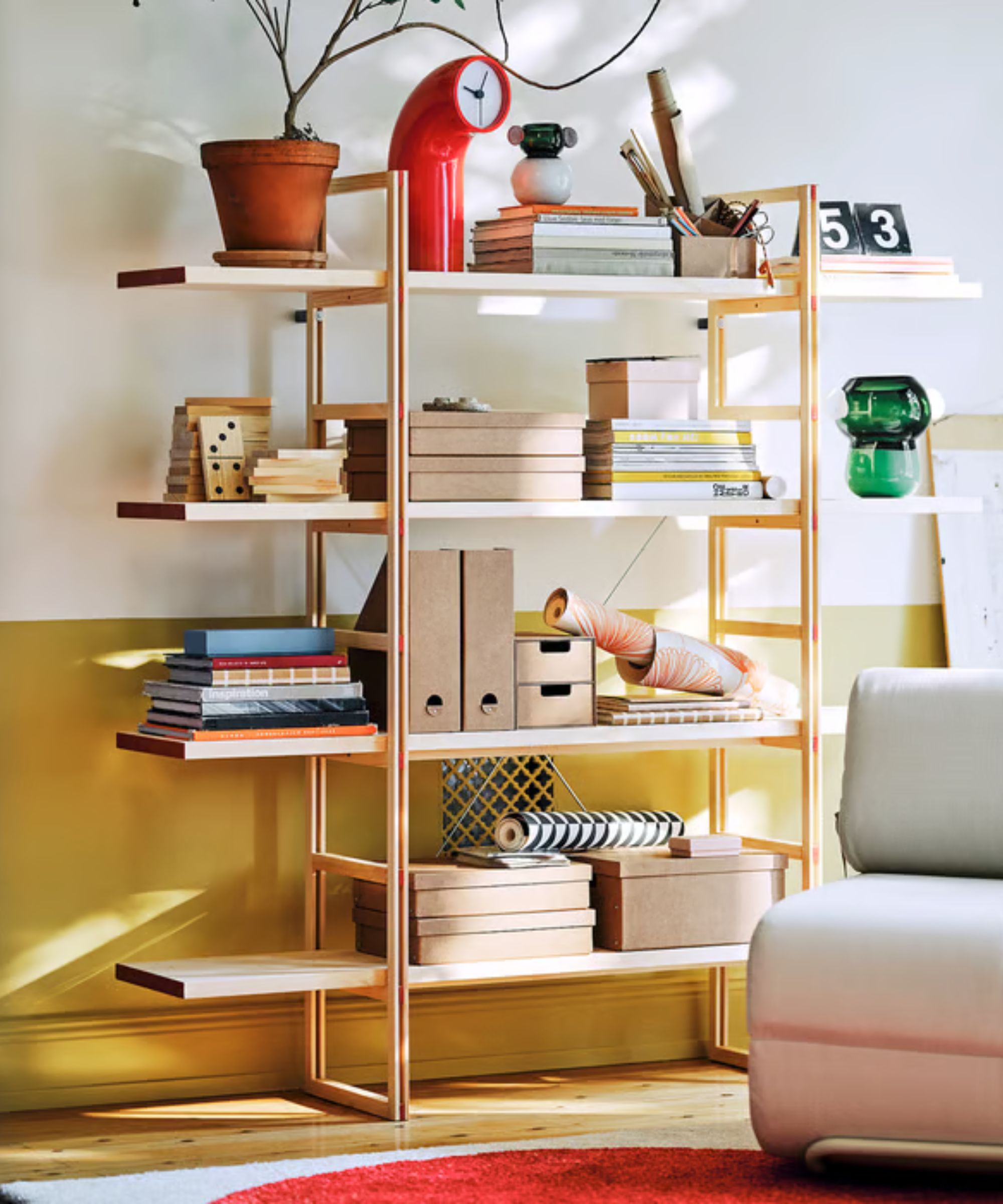 IKEA PS shelving unit decorated with objects, a clock, lots of stacks of books, and files, in front of a yellow and white wall, with a wooden floor and red rug.